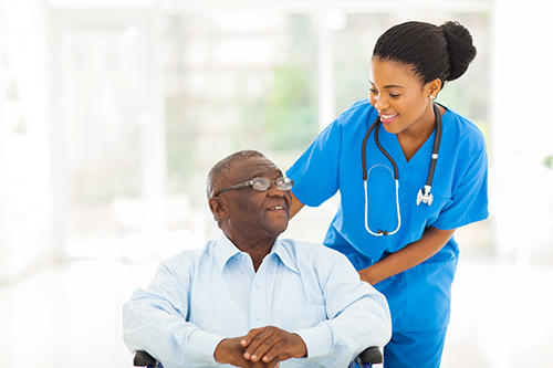 Young nurse in blue scrubs standing next to elderly man in wheelchair smiling at each other